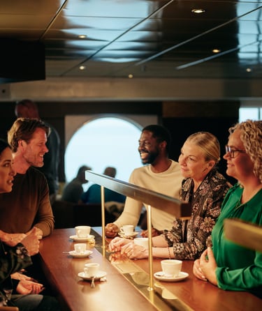 Gruppe af mennesker der sidder ved en disk og drikker kaffe, med store vinduer i baggrunden
