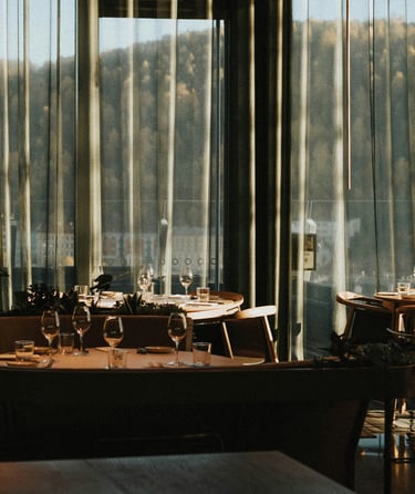 Elegant restaurantinteriør med opdækkede borde og udsigt til skov gennem store vinduer.