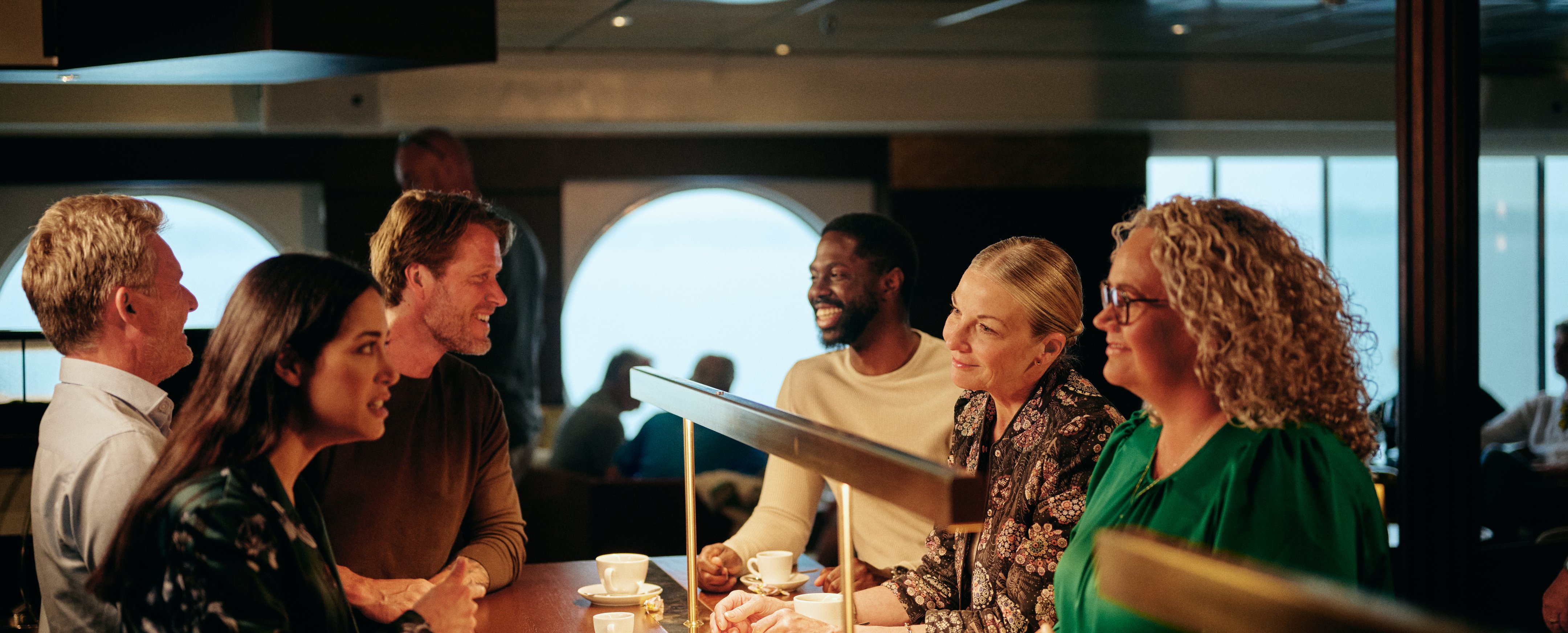 Gruppe af mennesker der sidder ved en disk og drikker kaffe, med store vinduer i baggrunden