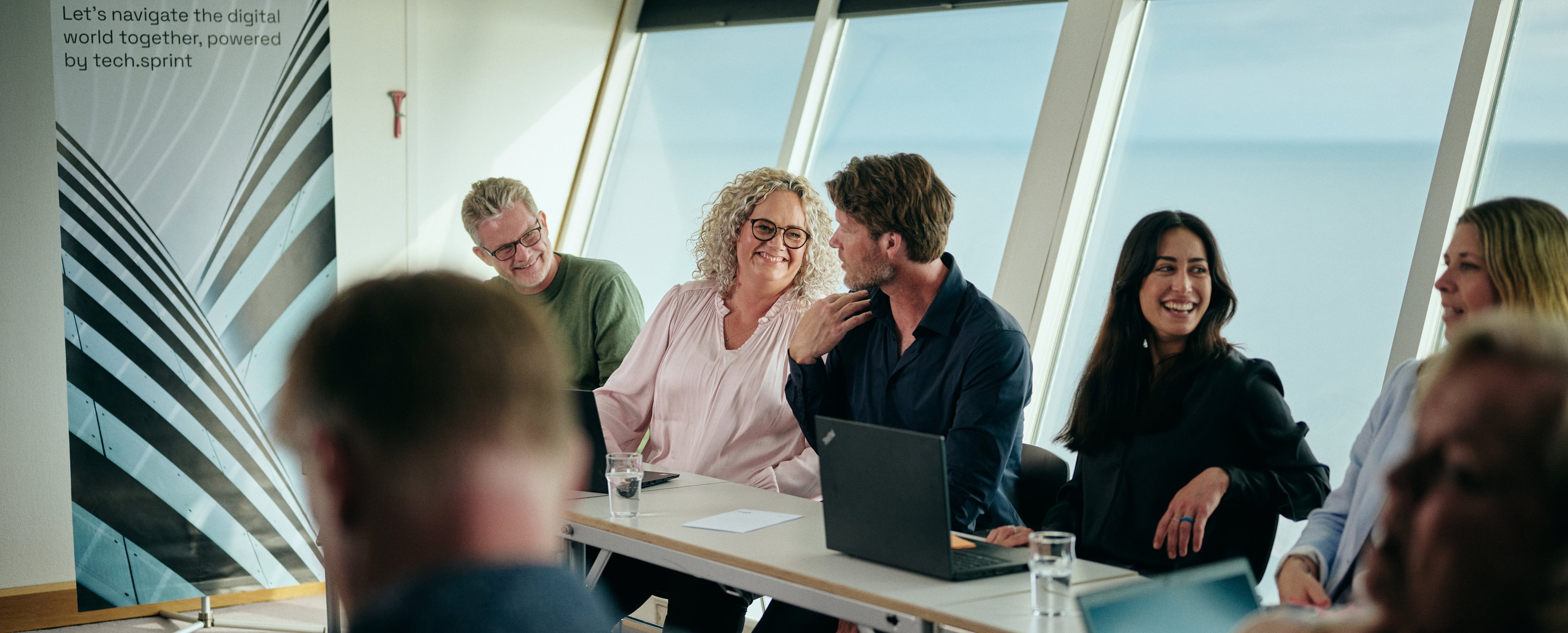 Møde med laptops ved vandudsigt, banner: "Welcome on board..."
