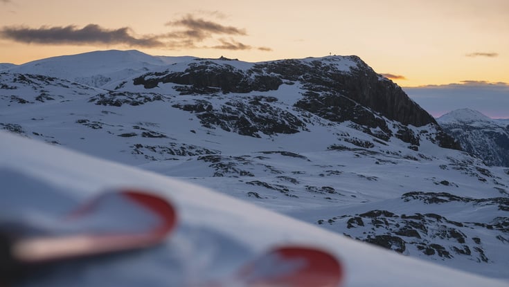 Snedækket bjerglandskab under solnedgang med skispidser i forgrunden.