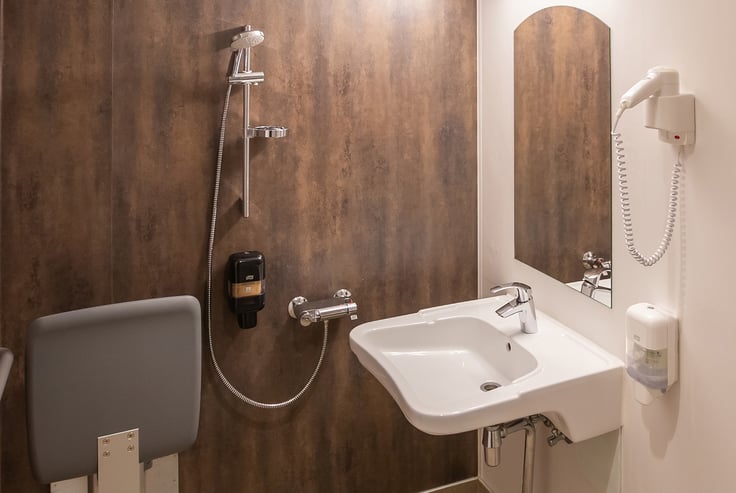 Handicapvenligt badeværelse med vask, bruser og sammenklappeligt sæde.