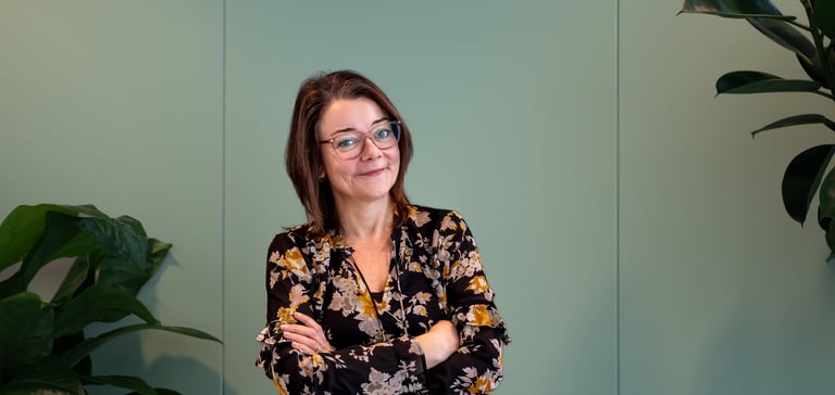 En selvsikker kvinde står med armene over kors foran en lysegrøn væg, omgivet af planter