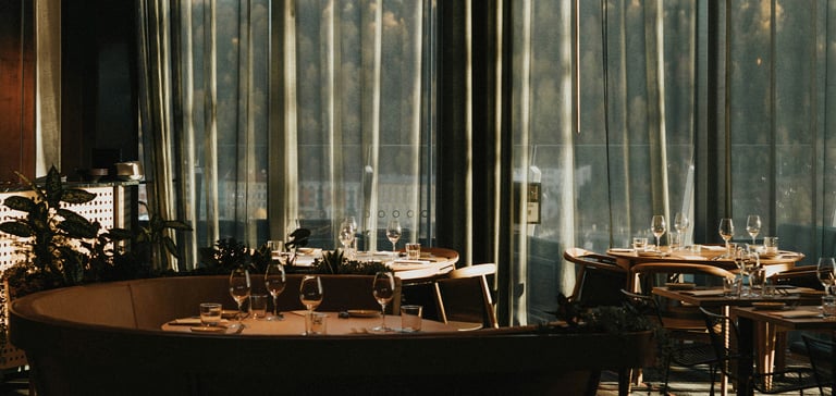 Elegant restaurantinteriør med opdækkede borde og udsigt til skov gennem store vinduer.