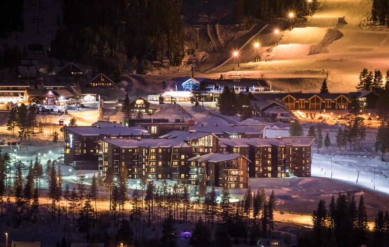 Natteudsigt over et snedækket feriested med oplyste bygninger og skiløjper.