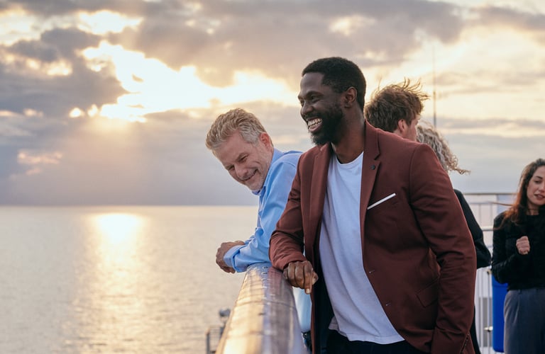 Fire personer, der står på en terrasse med havet og solnedgang i baggrunden.