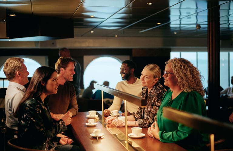 Gruppe af mennesker der sidder ved en disk og drikker kaffe, med store vinduer i baggrunden