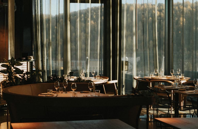 Elegant restaurantinteriør med opdækkede borde og udsigt til skov gennem store vinduer.